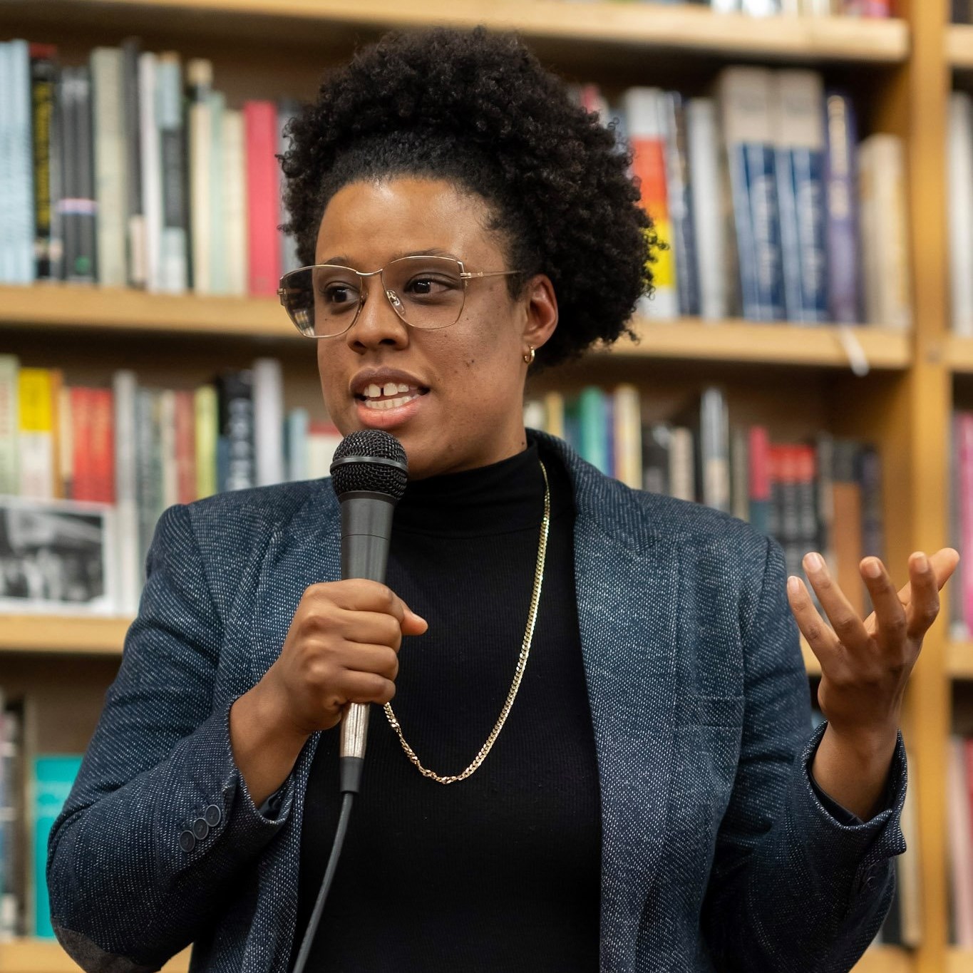 A color photo of Ejeris Dixon, speaking into a microphone. She has short hair, glasses, and is wearing a blue blazer with a black shirt underneath. She is standing in front of a bookshelf.