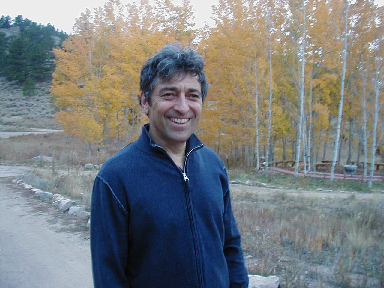 A color photo of Robert Gass, smiling at the camera. He has short hair, and is wearing a blue sweather. He is standing on a path in the woods.