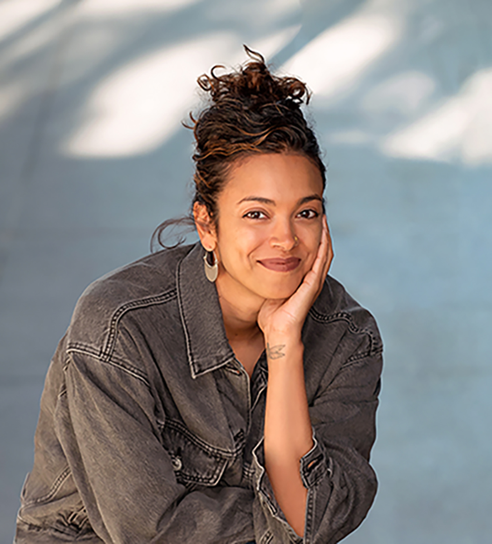 Vanessa Priya Daniel smiles at the camera with chin resting in left palm. Her brown hair is loosely up and she is wearing gold hoop earrings and a gray denim shirt.